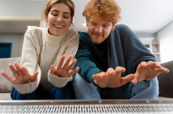 Alt text: Pár sedí pohodlne na pohovke a zohrieva si ruky pri radiátore.